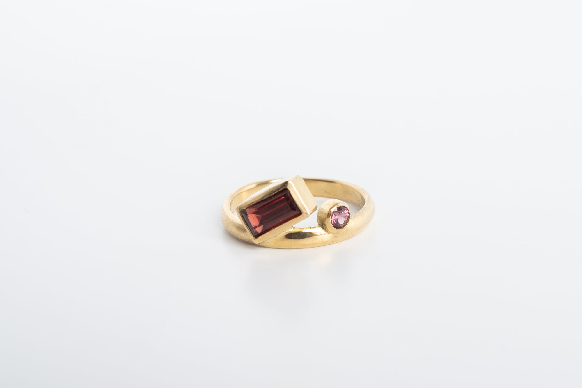 Gold ring with baguette cut garnet bezel set at a 45 degree angle to the band and a small 3mm round sapphire bezel set next to it, offset to sit above the ring band